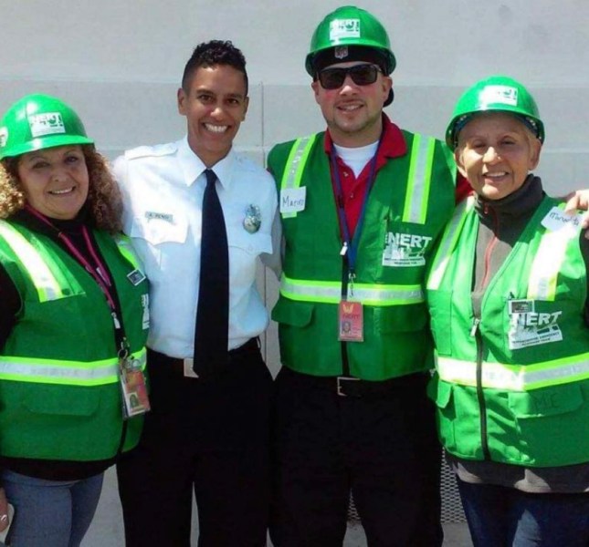A group of four individuals, including a uniformed firefighter and three volunteers, wearing green NERT safety vests and hard hats, pose together at an emergency preparedness training event.